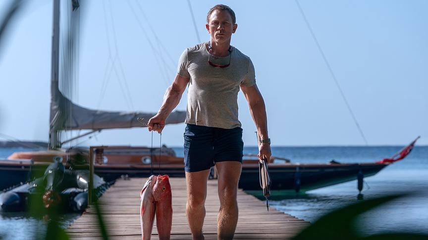 Ein Mann mit Fischen in der einen Hand und einer Harpune in der anderen geht einen Steg entlang, im Hintergrund ein Boot im Wasser