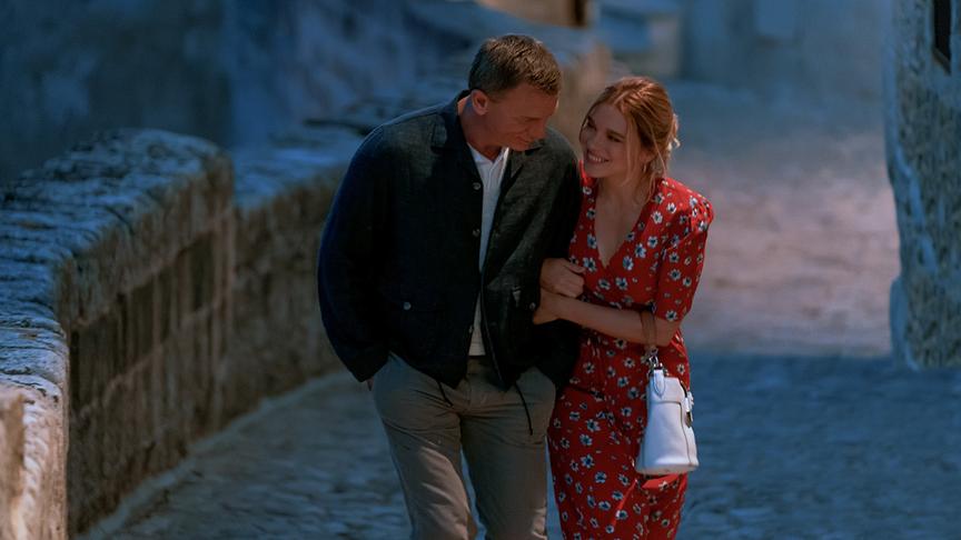 Ein Mann und eine Frau gehen eingehakt und lächeln eine Gasse mit Kopfsteinpflaster entlang.