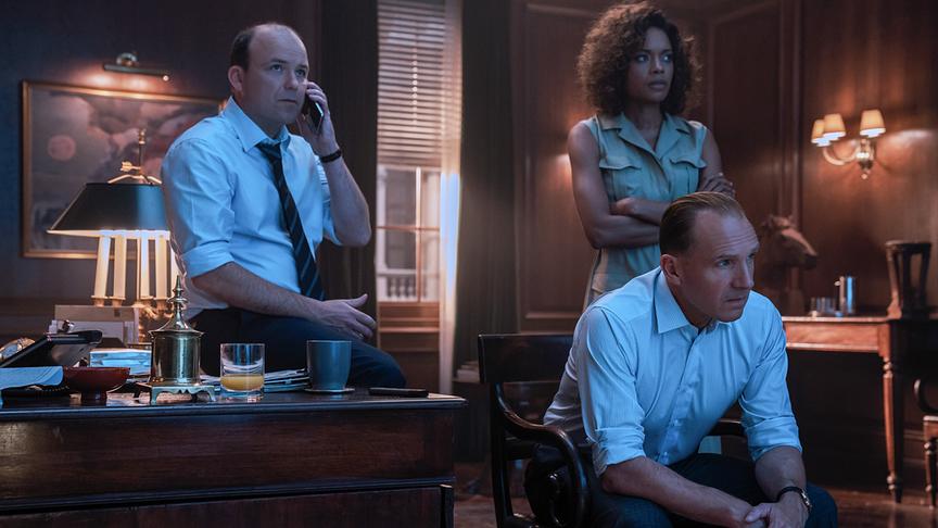 Zwei Männer und eine Frau in Business-Kleidung in einem Büro blicken gespannt auf etwas (wahrscheinlich einen Fernseher).