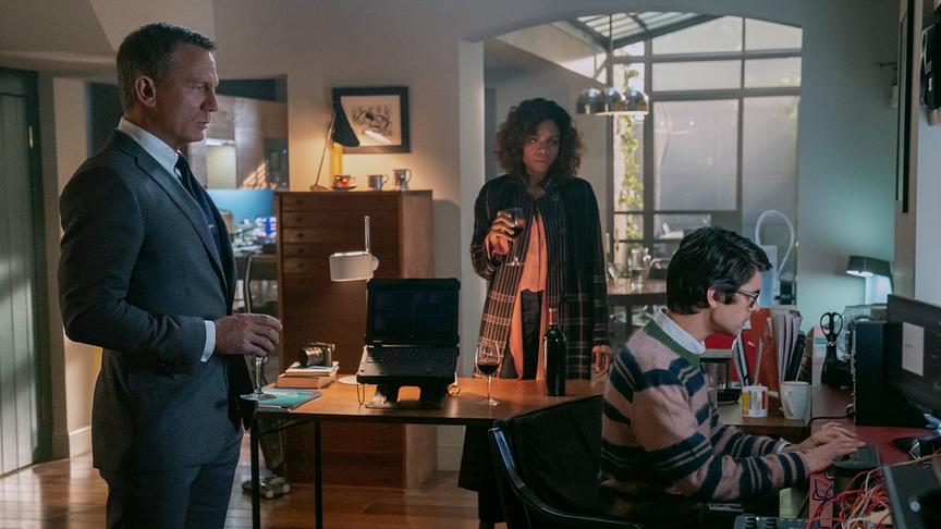 Ein Mann und eine Frau mit Weinglas in einer Wohnung, vor ihnen tippt ein Mann am Schreibtisch am Computer.