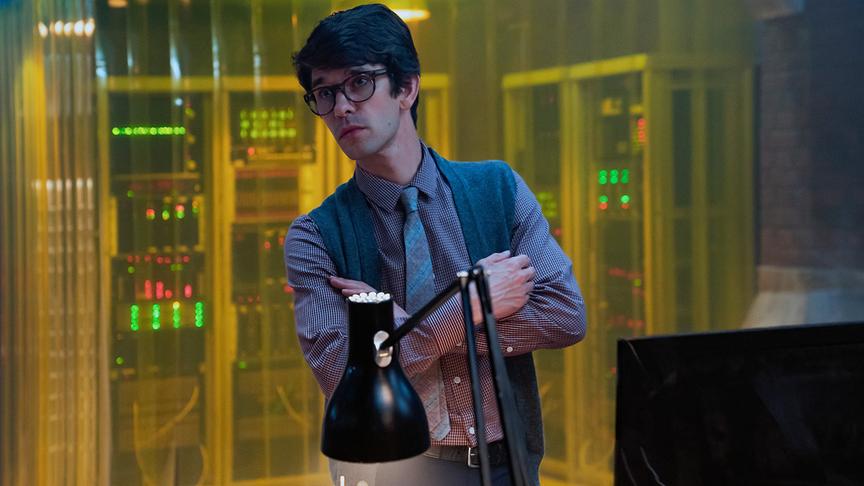 Ein Mann mit Brille und verschränkten Armen an einem Schreibtisch stehend, hinter ihm technische Geräte