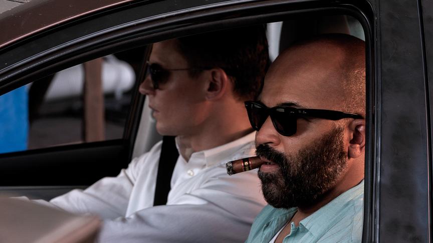 Zwei Männer mit Sonnenbrillen in einem Auto, einer davon raucht eine Zigarre