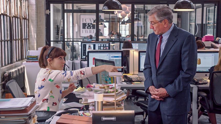 Eine Frau an einem vollbeladenen Schreibtisch und mit Headset weist einen Mann mit ausgestrecktem Zeigefinder an