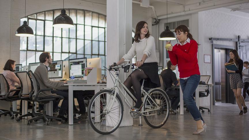 Eine Frau fährt mit dem Fahrrad durch ein Büro, neben ihr rennt eine andere Frau mit Notizblock und Stift in der Hand