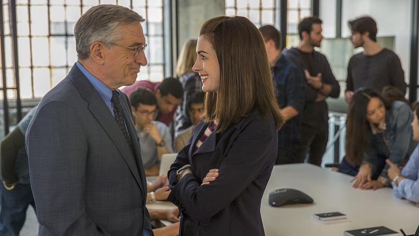 Mann und Frau stehen sich lächelnd gegenüber, im Hintergrund mehrere Personen an einem Tisch