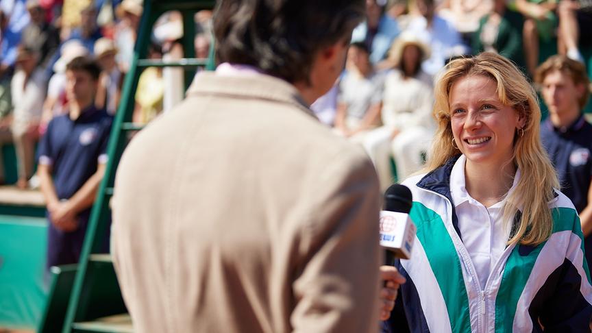 © Foto: ORF/Bravado Media/MarVista Entertainment/Umberto Poto Eine Frau steht gegenüber von einem Mann mit Mikrophon in einem Tennisstadion mit Publikum.