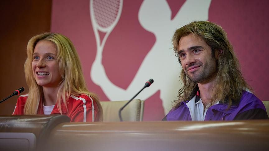 Eine Frau und ein Mann an einem Tisch mit Mikrofone. Im Hintergrund eine Wand mit weißer Tennisspielershilouette.