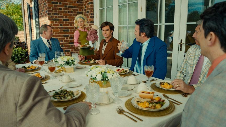 Um den gedeckten Tisch herum sitzen mehrere Männer im Anzug, während eine Frau mit Kind auf dem Arm einen Teller serviert.