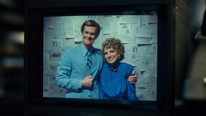 Ein Fernseher in dem eine Frau und ein Mann eng nebeneinander stehen vor einem Hintergrund aus aufgehängten Zeitungsartikeln.