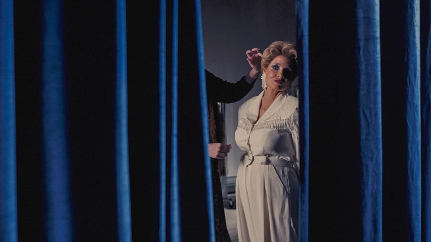 Eine Frau ist durch den Spalt eines blauen, schweren Vorhangs zu sehen. 