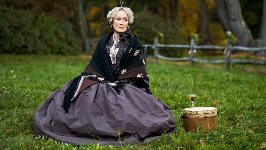 Eine ältere Dame sitzt im Gras, neben ihr ein rundes Gepäckstück auf dem ein Getränk in Glas steht.