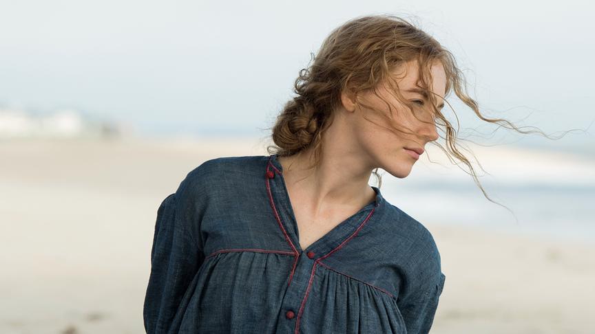 Ein Mädchen steht mit hinter dem Rücken verschränkten Armen am Strand, ihre Haare wehen in ihre Blickrichtung nach rechts.