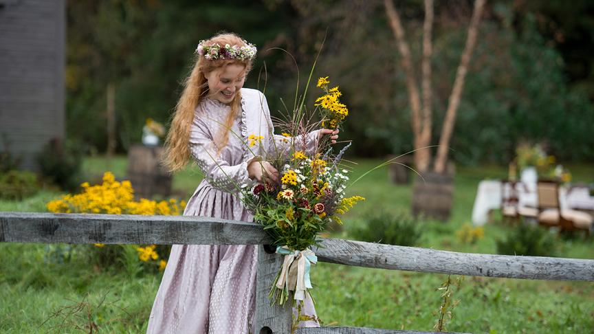 Ein Mädchen mit Blumenkranz im Haar dekoriert einen Zaunpfahl mit einem Blumenstrauß.