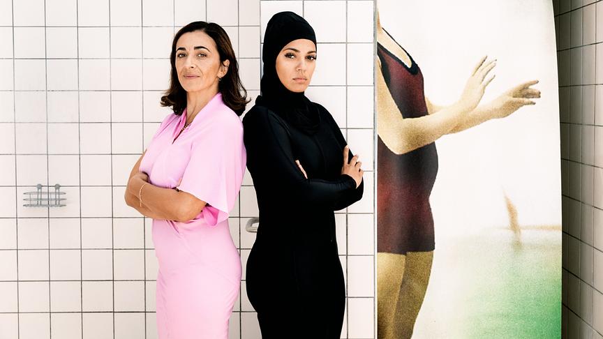Zwei Frauen stehen Rücken an Rücken, eine in schwarz bekleidet und mit Kopftuch, die andere in rose Kleidung
