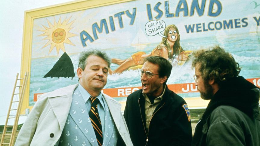 Drei Männer nebeneinander, einer scheint auf den Mann im Anzug einzureden; im Hintergrund ein Plakat mit einer Frau, die von einem Hai verfolgt wird