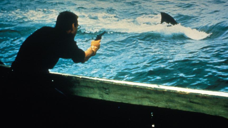 Mann zielt mit einer Pistole auf eine aus dem Wasser ragende Haiflosse