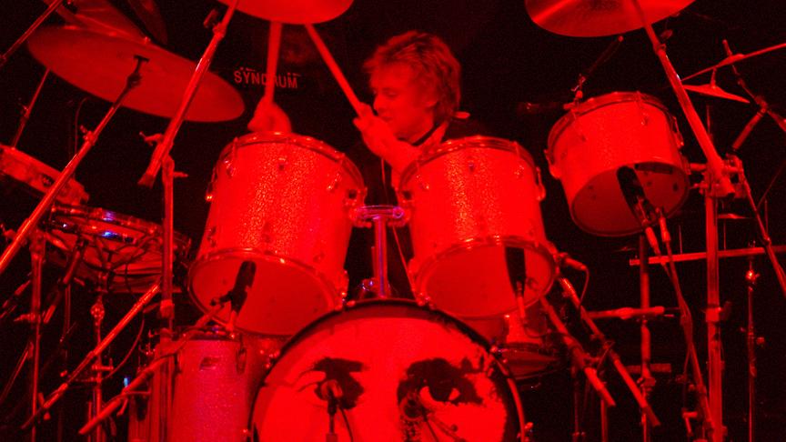 Roger Taylor spielt am Schlagzeug und ist in rotes Licht gehüllt.