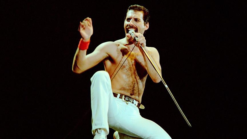 Freddy Mercury oberkörperfrei in einer Pose kniend und dabei in ein Mikrofon singend.