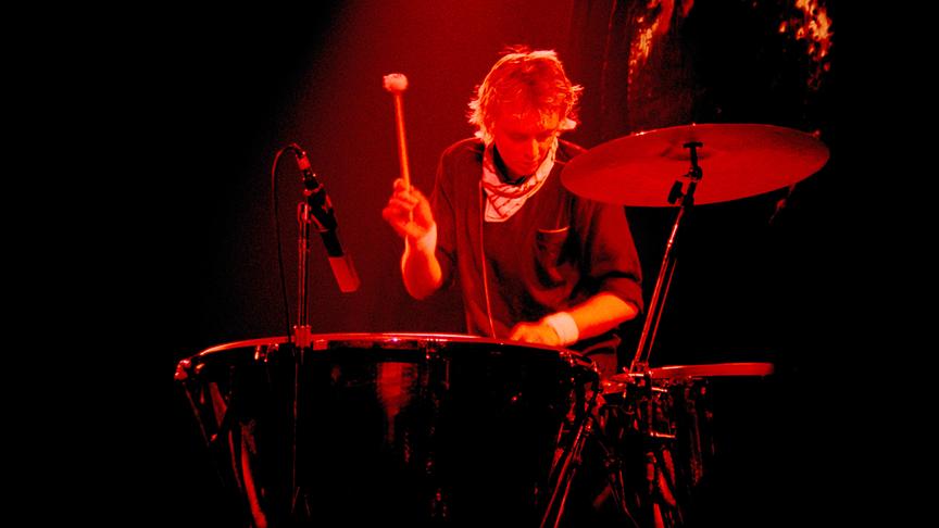 Roger Taylor am Schlagzeug spielend und in rotes Licht getaucht.