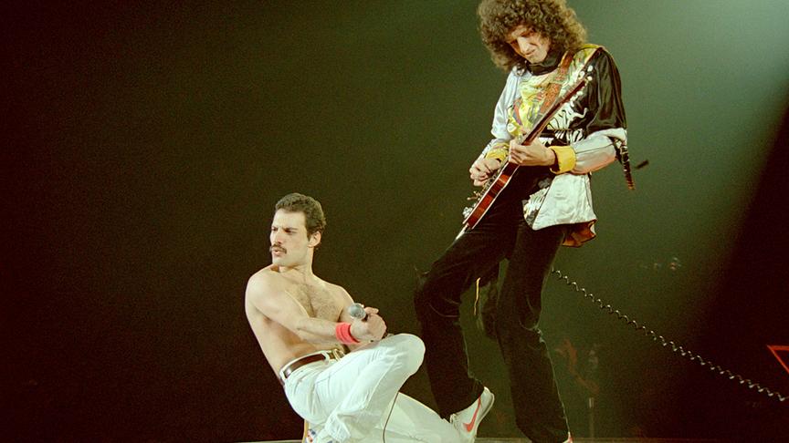 Freddy Mercury oberkörperfrei in einer Pose am Boden kniend, danebem Brian May stehend und Gitarre spielend.