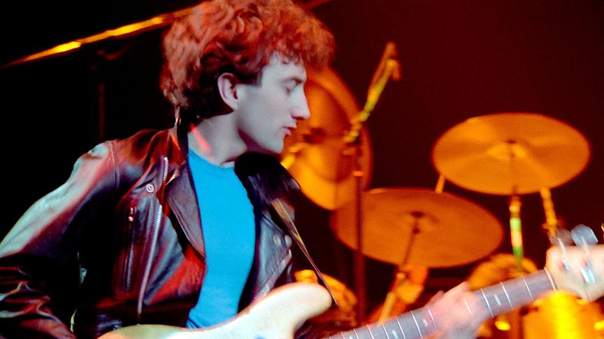 John Deacon in Lederjacke und Gitarre spielend.