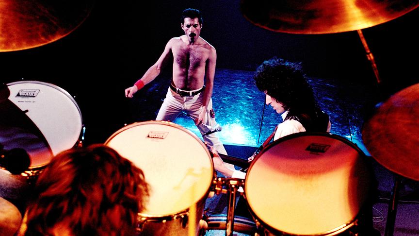 Aufnahme von einem erhöhten Podest, auf dem ein Schlagzeug steht, hinunter auf die Mitglieder von Queen. Freddy Mercury steht oberkörperfrei und dem Schlagzeug zugewandt auf der Bühne.