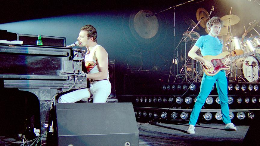 Freddie Mercury im weißen Outfit an einem Piano und am Mikrofon. Danben ein anderes Bandmitglied an der Gitarre und in blauem Outfit.