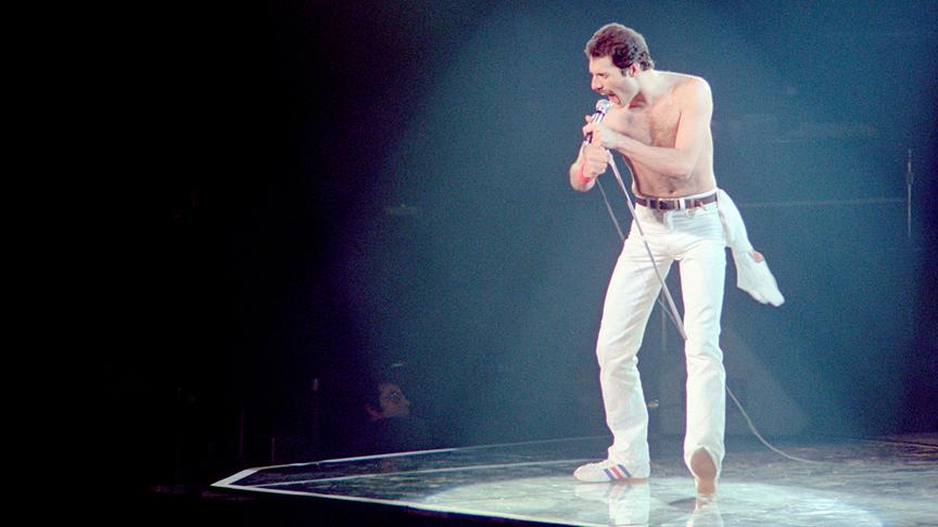 Freddie Mercury oberkörperfrei in weißer Hose auf einer Bühne im Rampenlicht.