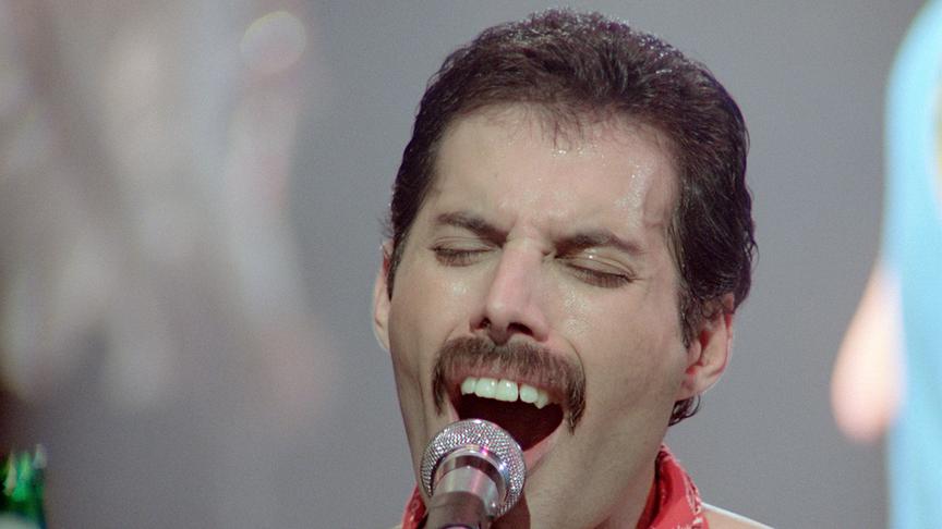 Freddie Mercury singt inbrünstig in ein Mikrofon. Er trägt einen Schnauzbart und und ein rotes Halstuch.