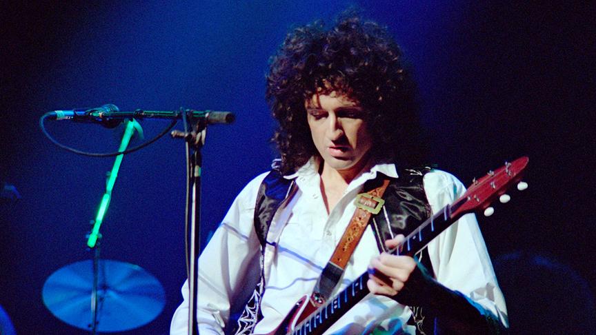 Brian May an der Gitarre und hinter einem Mikrofon bei blauer Beleuchtung auf einer Bühne.