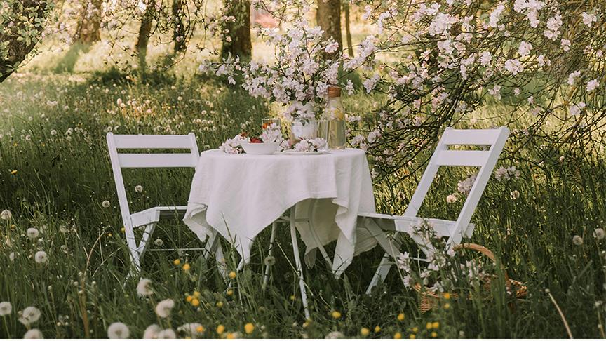 Ein weißer Tisch gedeckt mit Tischdecke, Blumen, Karaffe etc in einer blühenden hohen Wiese umgeben von blühenden Bäumen.