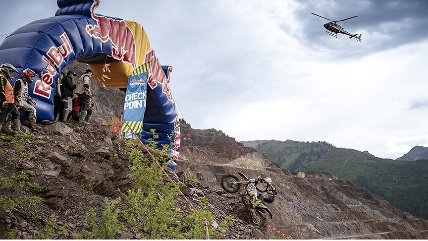 Motocrossfahrer fährt einen steilen Berg hinauf zum RedBull Checkpoint. Im Hintergrund der Erzberg und in der Luft ein Helikopter.