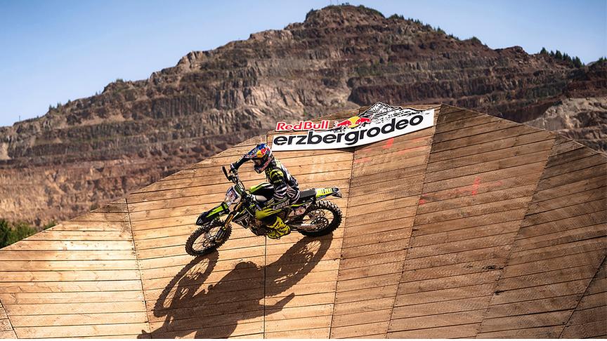Motocrossfahrer auf einer Rampe mit Red Bull Erzbergrodeo Logo. Im Hintergrund der Erzberg.