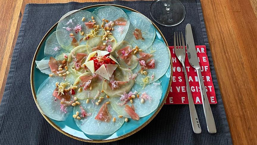 Ein gedeckter Tisch mit gefülltem Weinglas und einem Teller mit kunstvoll angerichtetem Prosciutto mit Käse.