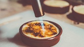 Eine Hand hält einen Küchengasbrenner und schmilzt den braunen Zucker auf einer Crème brûlée in einer Keramikform, sodass eine goldbraune Kruste entsteht.