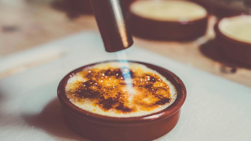 Eine Hand hält einen Küchengasbrenner und schmilzt den braunen Zucker auf einer Crème brûlée in einer Keramikform, sodass eine goldbraune Kruste entsteht.