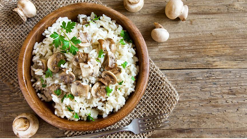 Holzschüssel mit Pilzrisotto, daneben eine Gabel und ein paar Champignons