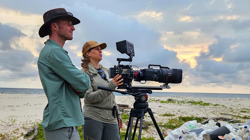 Filmteam in Outdoorkleidung steht mit einem Kamerastativ am Strand. 