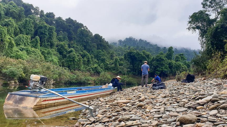 Drei Menschen sind auf einem steinigen Ufer neben ihnen ist ein Boot im Fluss.