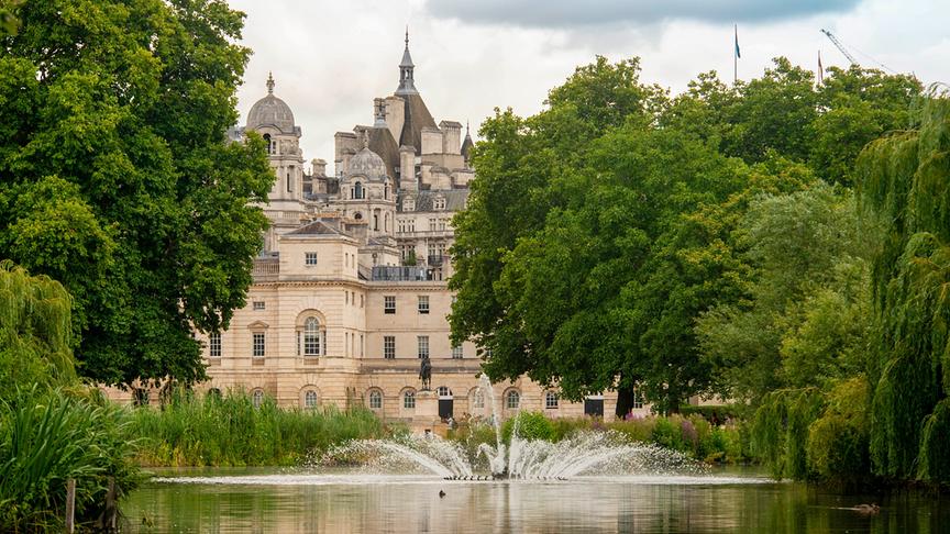 Ein Park mit Bäumen und einem Gewässer mit Springbrunnen vor einem schlossähnlichen Gebäude. 