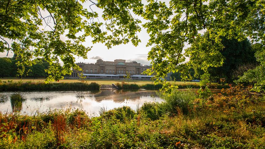Grüne Landschaft mit Gewässer, Bäumen und Wiese; im Hintergrund der Buckingham Palace