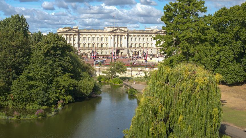 Im Hintergrund der Buckingham Palace, im Vordergrund ein Park mit Bäumen und einem Gewässer.