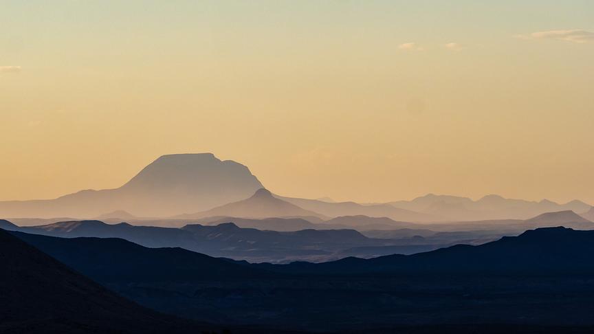 Weite Landschaftsaufnahme von mehreren, hintereinander liegenden Bergketten in orange-gelblichen Dunst.