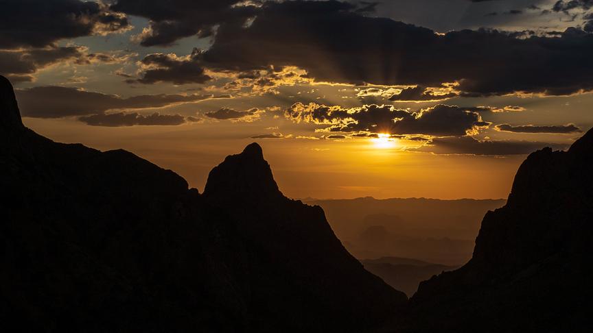 Silhouetten von Bergen bei Dämmerung. Die Sonne scheint warm unter den Wolken am Himmel hindurch.