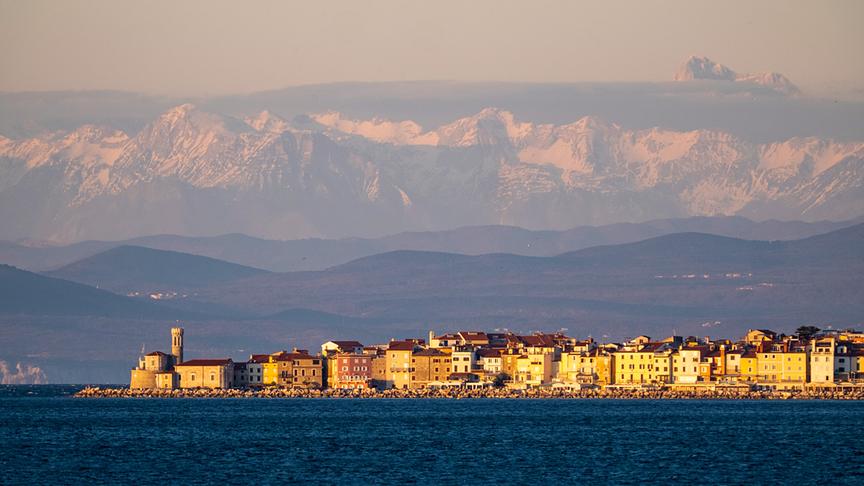 Im Vordergrund erstrecke sich die bebaute Küstensiedlung, während sich im Hintergrund zunächst Hügel und dann hohe Berge erstrecken.