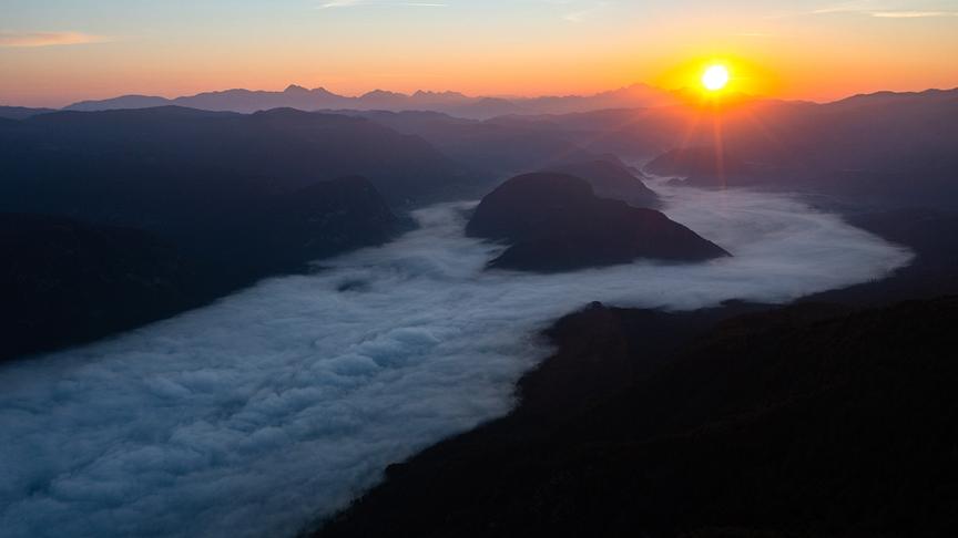 Die Nebeldecke hängt in Tal fest, während darüber die tiefstehende Sonne über dem Bergpanorama steht.