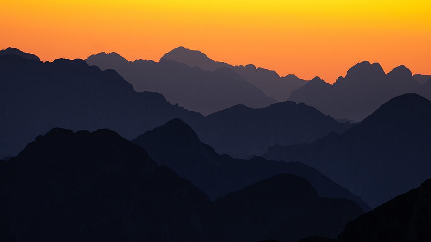 Durch die orange-leuchtende Sonne sind die einzelnen, nebeneinander verlaufenden Bergketten nur als Umrisse erkennbar.