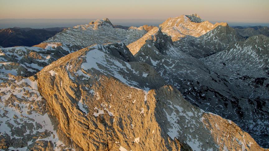 Eine karge, felsige Gebirgskette, die leicht von Schnee bedeckt ist und von warmen Licht seitlich angestrahlt wird.
