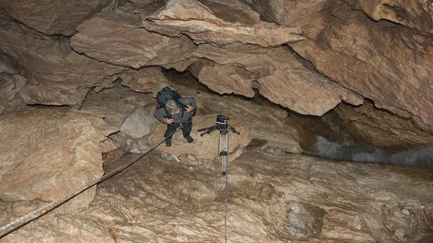 Eine Felshöhle von oben herab fotografiert. Zwei Seile führen hinunter, unten steht eine Person mit Rucksack.