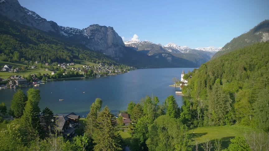 Der Grundlsee zwischen hohen Bergen und bewaldeten grünen Flächen liegend unter blauem Himmel.